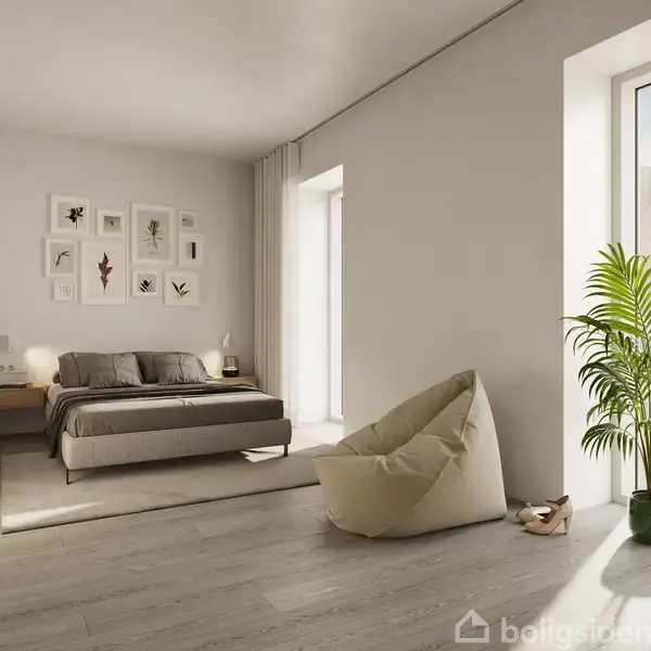 En dobbeltseng står i et minimalistisk soveværelse med lysgrå vægge og træmøbler. Ved siden af sengen stands en lænestol og en plante ved store vinduer, der lukker lys ind.