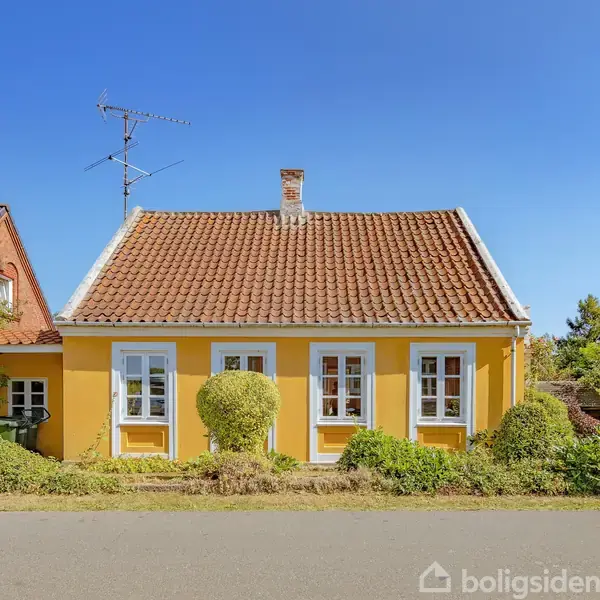 Gult hus med hvidrammede vinduer og rødt tegltag står stille under blå himmel, omkranset af frodig have med buske og træer i et villakvarter.