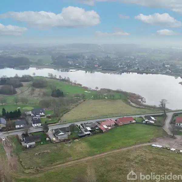 En droneskud viser et bakket landskab med spredte huse langs en vej, mens en stor sø strækker sig i baggrunden under en delvist skyet himmel.