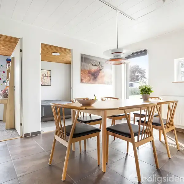 Et spisebord med fem stole står i et lyst køkken. En lampe hænger over bordet. Store vinduer og planter skaber en luftig atmosfære.