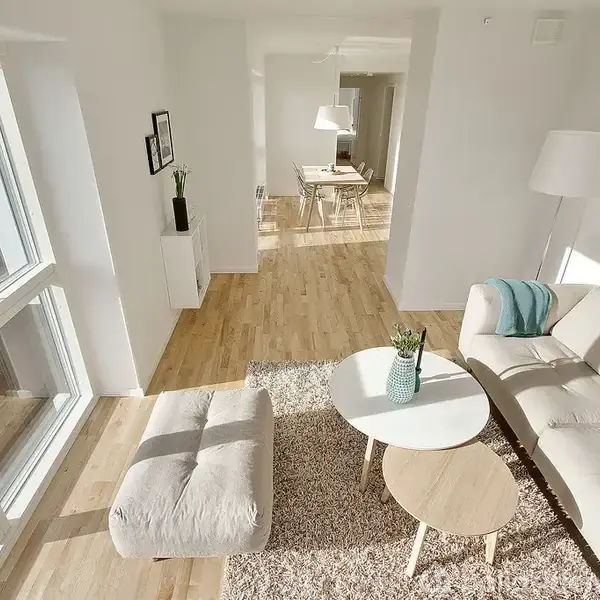 En lys stue med en beige sofa og to runde borde placeret på et tæppe. En stor vinduesparti giver udsigt til bylandskab. Dårlig belysning afslører også en bordlampe og spiseområde i baggrunden.