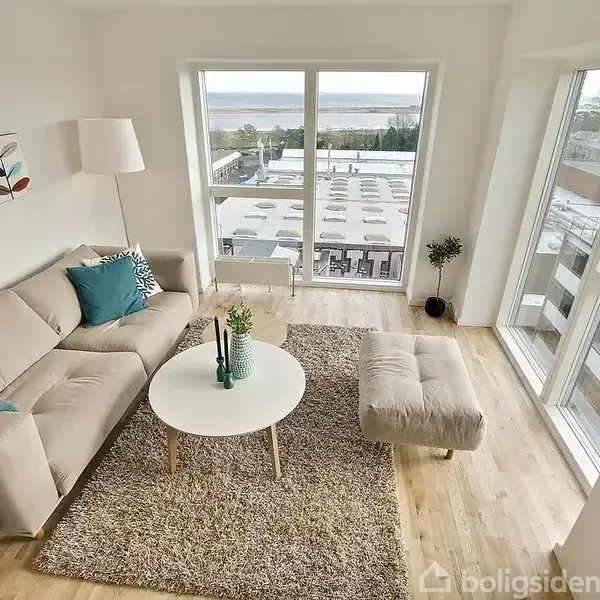 En lys stue med en beige sofa fyldt med puder og en plaid, et rundt sofabord med dekorationer, og et stort vindue med udsigt over et bylandskab og vand i baggrunden.