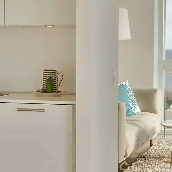 Et lyst køkken med en hvid håndvask og et stribet krus. Til højre ses en sofa med blå puder og et sofabord med stearinlys. Udsigt til havet udenfor.
