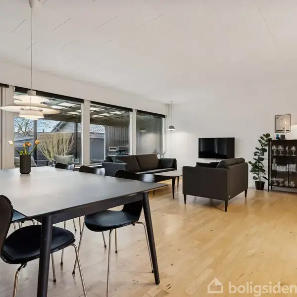 Spisebord med sorte stole står i en lys stue. Sofaer er placeret foran et TV. Store vinduer åbner ud til en terrasse. En reol står i hjørnet.