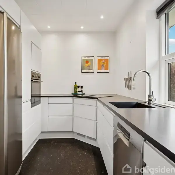 Et moderne køkken med hvide skabe og stål-apparater. To billeder hænger på væggen over en sort bordplade med en vask. Store vinduer giver udsigt til en busk.