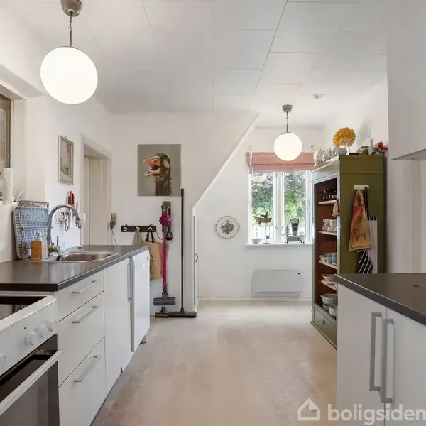Et lyst køkken med hvide skabe og sorte bordplader strækker sig langs to vægge. Midt i rummet hænger runde loftlamper. Et vindue for enden giver naturligt lys.
