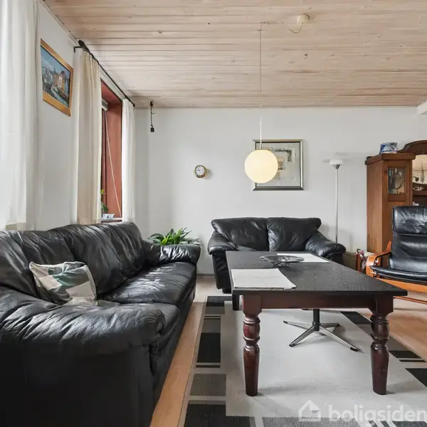 Sort lædermøblement står i stue med trægulv og loft. En sofa og en lænestol står omkring et sofabord. I baggrunden ses et skab og en lampe. Vinduer giver naturligt lys.