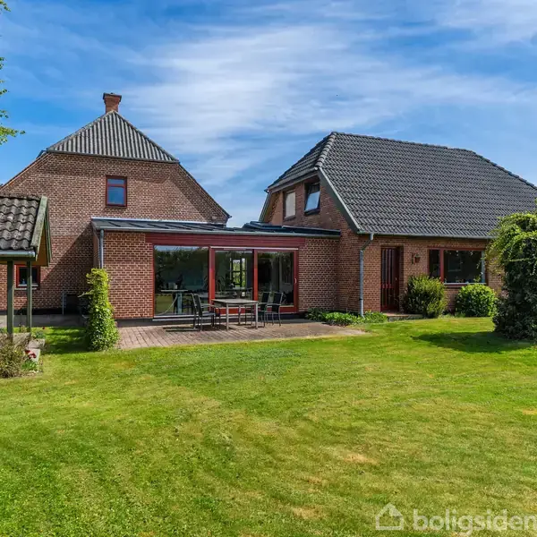 Rødt murstenshus med sort tag står stille i en velplejet have. En terrasse med havemøbler ses foran huset, og en blå himmel danner baggrund.