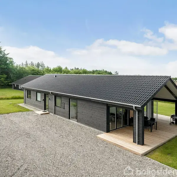Sort træhus står stille med en terrasse, omgivet af grønne græsplæner og træer. En stenbelagt indkørsel fører op til huset, der ligger i et naturskønt område.