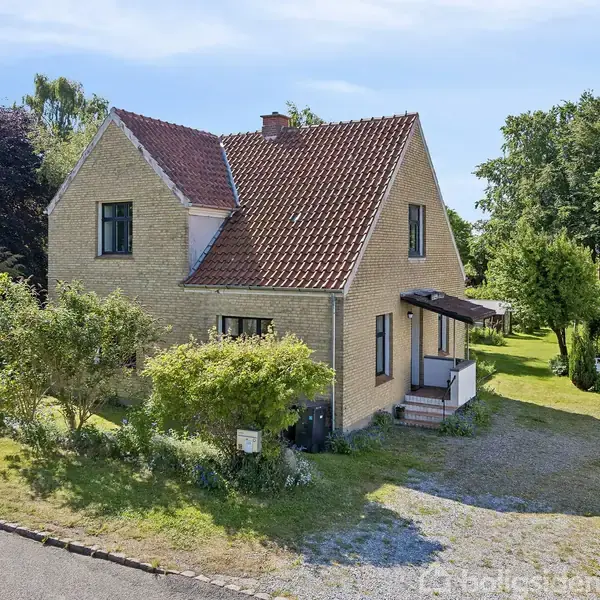 Et gult murstenshus med rødt tegltag står i en frodig have. En grusvej fører til en carport med en bil. Omkring huset er der flere træer og buske.