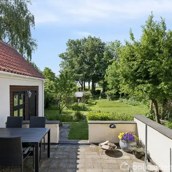 Terrasse med havemøbler fører ud til en velplejet have med træer og buske. Bygningen til venstre har et rødt tegltag. Himlen er blå og solbeskinnet.