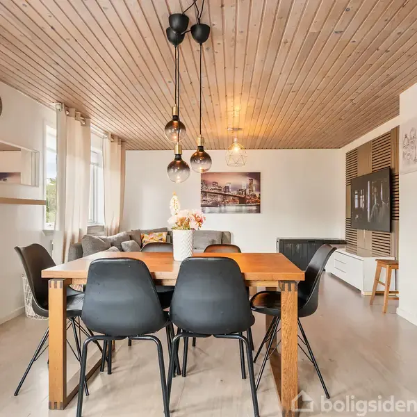 Spisebord med seks sorte stole står under hængende lamper i en lys stue. I baggrunden er en sofa med puder, flere vinduer og vægdekorationer. Karton med teksten "Tineke & And...".