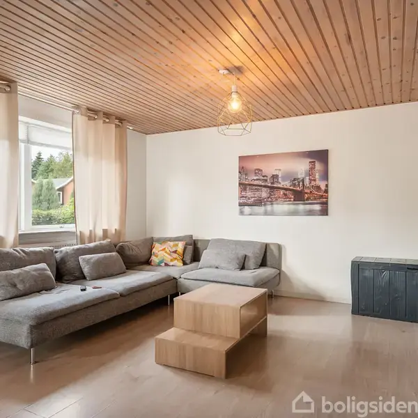Gråt hjørnesofa står i stue med lyse trægulve og træbeklædt loft. Et billede hænger på den hvide væg, og et sofabord står foran sofaen. Vinduer med gardiner giver naturligt lys.