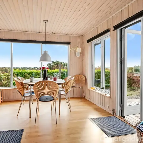 Spisebord med seks stole står i et rum med store vinduer, der giver udsigt til en terrasse og grønne marker. En stol og en lænestol ses også i rummet.