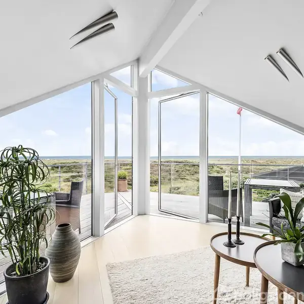 Et lyst rum har store vinduer med udsigt over en terrasse og en sandklit-landskab. En sofa med puder og et lille rundt bord står i rummet. En stor plante står ved vinduet.