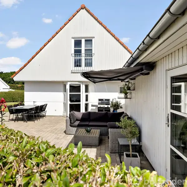 Hvidt hus med rødt tag står i en have. Havemøbler og en grill er på terrassen, omgivet af grønne buske. Solskin og blå himmel danner en lys baggrund.