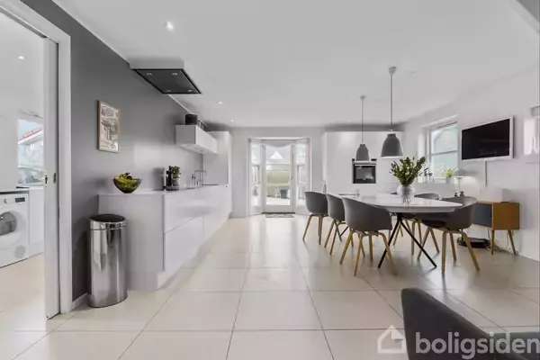 Et lyst, moderne køkken-alrum med spisebord og stole. Køkkenet har hvide skabe og en sølvfarvet skraldespand til venstre. Store vinduer og en dobbeltdør giver udsigt til haven.