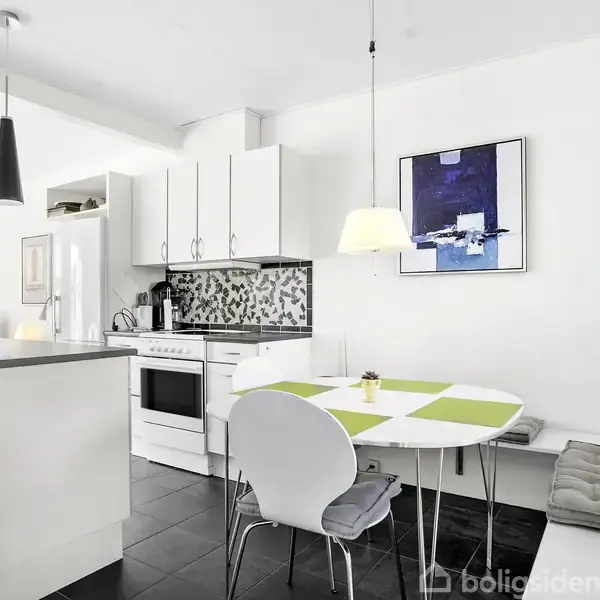 Et moderne køkken med hvidt interiør og sort-hvid mosaikflisevæg. Der er et rundt spisebord med grøn-hvid dug og to stole. Over bordet hænger en lampe. Billedet har et pænt indrettet miljø.
