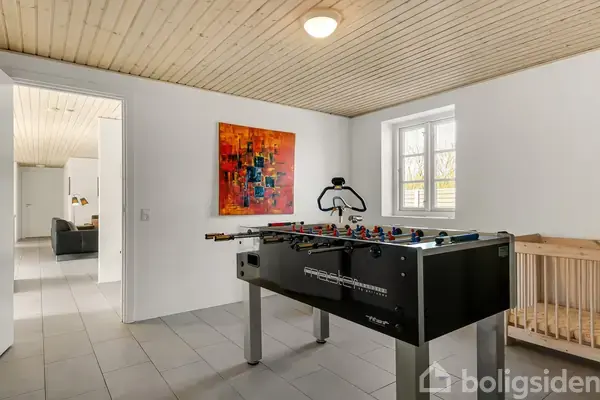 Bordfodboldbord står i et lyst rum med hvidmalede vægge og træloft. Der er et abstrakt maleri på væggen, et vindue til højre og en døråbning til venstre.