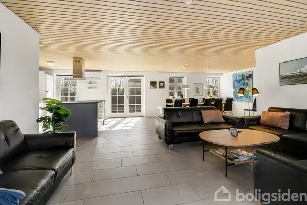 Sort lædersofa står i moderne stue med åbent køkken. Rummeligt med stort vinduesparti, lyst trægulv og spisebord med stole. Lyse vægge og dekorationer giver en hyggelig atmosfære.