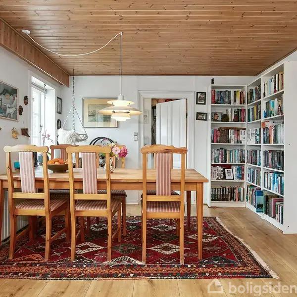 Spisebord med fire stole står på et orientalsk tæppe. En loftslampe hænger over bordet. Væghylde og bogreol med mange bøger fylder rummet, som har et lyst trægulv.