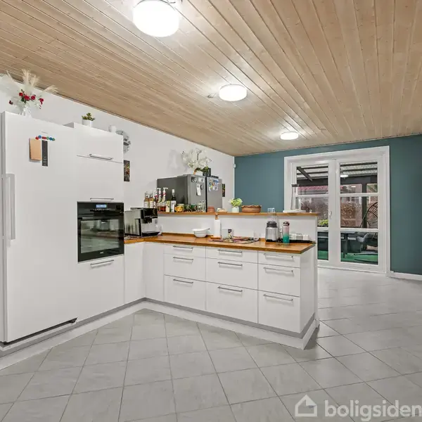 Et moderne køkken med hvide skabe og træbordplader, er centralt i billedet. Køkkenet har hvidevarer og dekoration. En dør fører ud til en terrasse. Hvidt loft og grøn væg som kontrast.