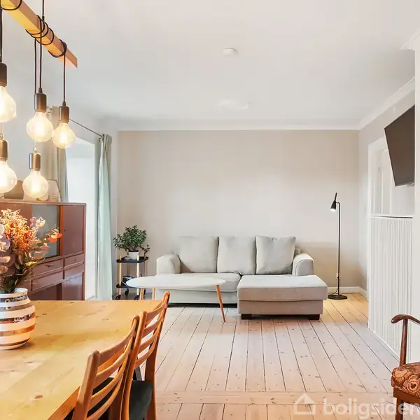 En stue med en grå sofa står mod en lys væg. Et træbord med seks stole og en vase med blomster står foran. Hængelamper lyser rummet op.