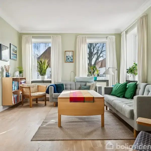 Stue indeholder en grå sofa med grønne puder, et træbord med farvestrålende dug, omgivet af store vinduer med hvide gardiner. Væggene har kunst og en bogreol står til venstre.