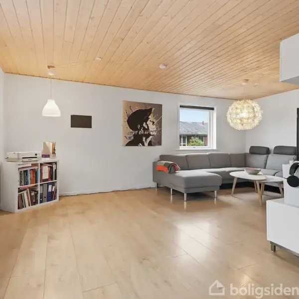 Grå hjørnesofa står i en moderne stue med lyst trægulv. Ved siden af er et rundt sofabord. Væggene er hvide med kunst. Store vinduer slipper lys ind.
