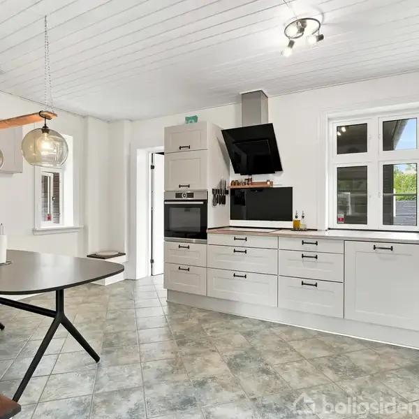 Et moderne, lyst køkken med hvide skabe og sort emhætte. Spisebord med hængende lamper ved siden af. Vinduerne giver udsigt til en grøn have.