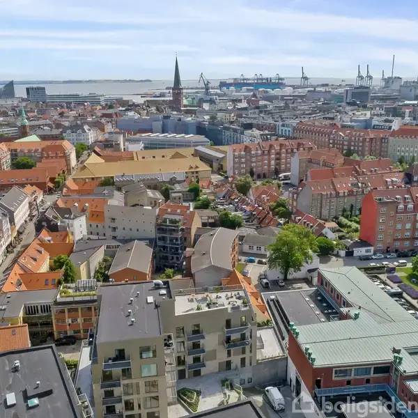 Bygninger med røde tage står stille, omgivet af et tætpakket bylandskab og en havn i det fjerne. Grønne områder bryder den urbane tæthed, og en kirketårn rejser sig blandt bygningerne.
