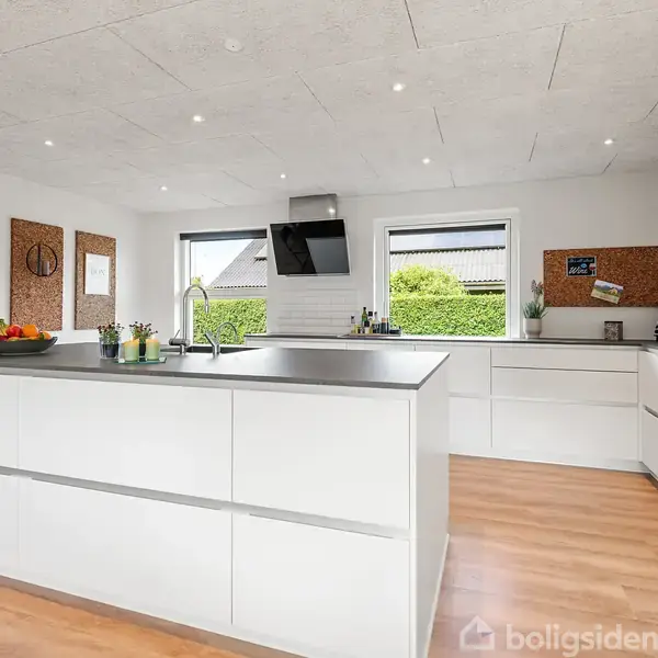 Køkkenø med moderne hvidt interiør, centreret i et lyst rum. Køkkenredskaber er placeret på bordet, mens vinduer i baggrunden giver udsigt til en grøn have.