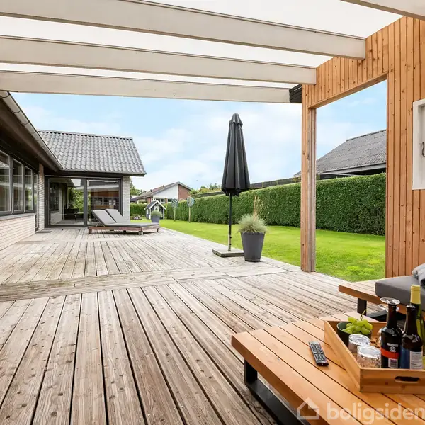 Terrasse med trædæk har to liggestole og et sort parasol. En sofa med et bord fyldt med vin og snacks står under en delvis overdækning. Græsplæne og hække omringer området.