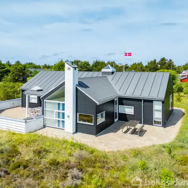 Sort, moderne sommerhus med dansk flag på taget, beliggende i grønne omgivelser med græs og buske. Huset har en terrasse med stole og bord samt et skorstensrør langs siden.