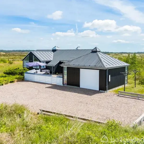Hus med mørkt tag og garage står på grusgrund, omgivet af åbent landskab. En dansk flagstang står til venstre, mens terrassen har parasol og borde.