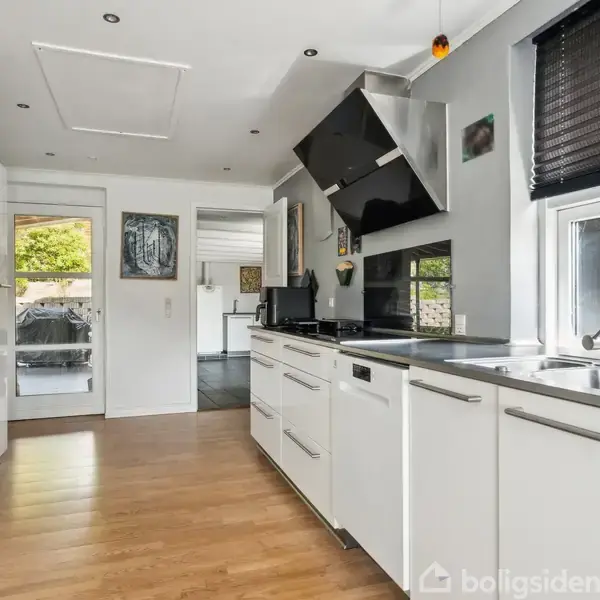 Et moderne køkken med hvide skabe, sorte apparater og en vask ved vinduet. Trægulve og en glasdør fører til en terrasse med grønne omgivelser.