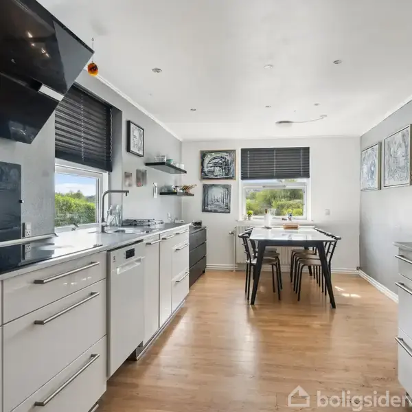 Køkken med hvide skabe og sort bordplade, udstyret med ovn og emhætte, i et lyst rum. I baggrunden står spisebord og stole ved et vindue med udsigt.