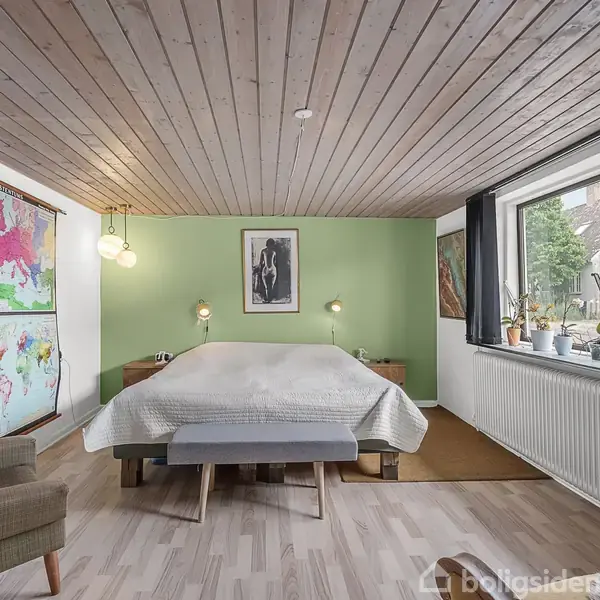 En spisestue med et stort træbord og fire stole. En kobberfarvet lampe hænger over bordet. Væggen har billeder og en grøn plante. Store vinduer lukker lys ind.