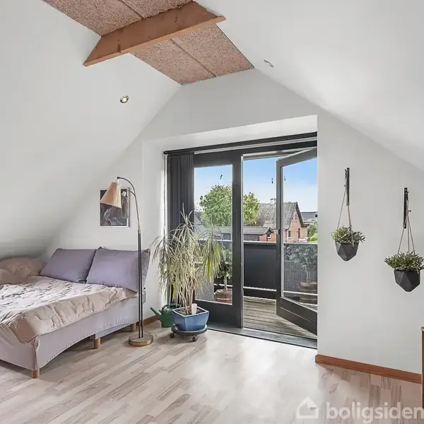 Soveværelse med en stor seng i midten, omkranset af natlamper. I højre side er der et stort vindue med udsigt og blomster, og på venstre væg hænger farverige kort.