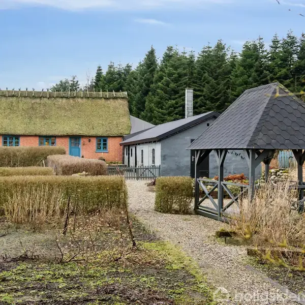 Bondegård med stråtag ligger stille omgivet af hække. En grussti fører til et lysthus i haven. Tætliggende skov markerer baggrunden under en blå himmel.