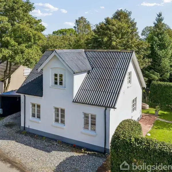 Hvidt, to-etagers hus med skråt tag står på en stille villavej, omgivet af velplejet grøn have og tæt hæk. Flere træer danner baggrund. Ingen synlig tekst.