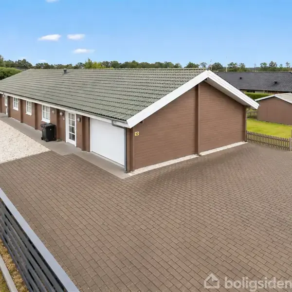En lang, brun murstensbungalow med grønt tag står i et boligområde. Den ligger ved en flisebelagt gårdsplads og er omgivet af en hæk.