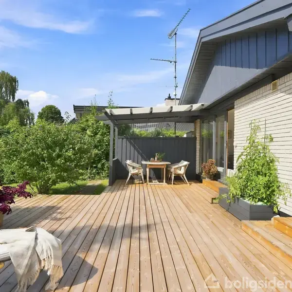 En terrasse med trægulv har havemøbler og planter i krukker. Terrassen er ved et hus med en grå og hvid facade. I baggrunden ses en frodig have og blå himmel.