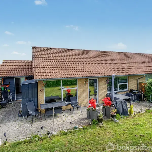Et enkelt enfamiliehus med rødt tegltag og gule murstensvægge. Foran huset findes en terrasse fyldt med havemøbler og blomsterkrukker, omgivet af en velplejet græsplæne.