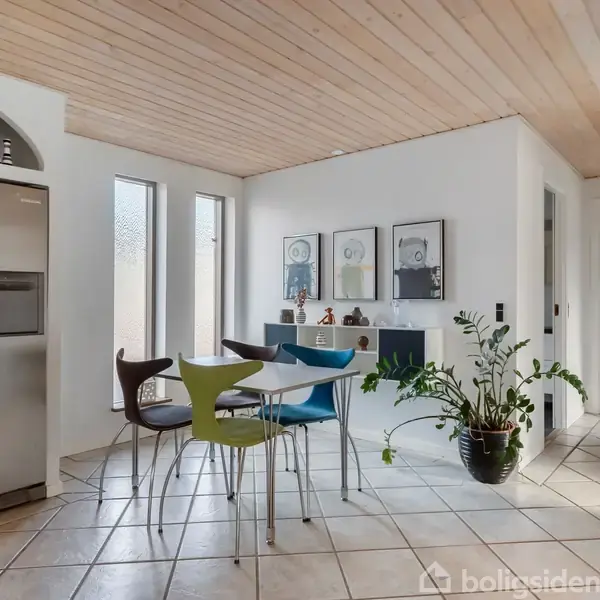 Et spisebord med stole står i et lyst køkken med flisegulv. Til venstre ses et stort køleskab, og til højre er der en grøn plante. Væggen har indrammede billeder.