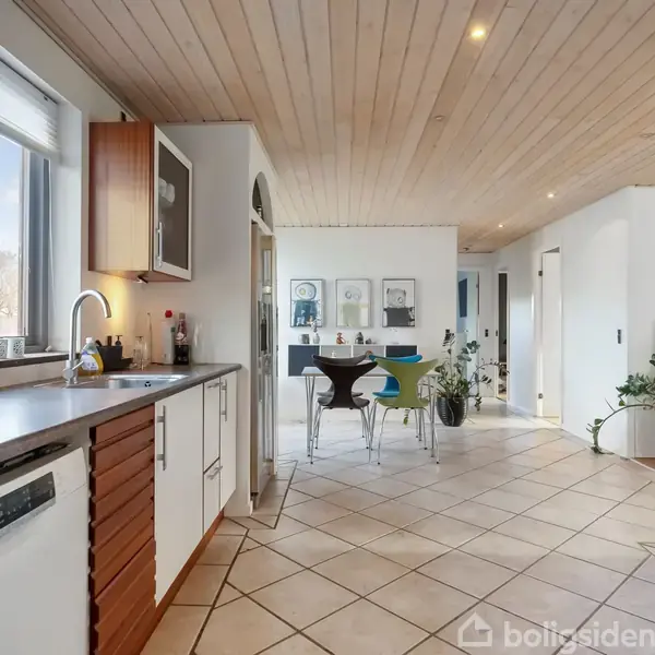 Køkken med hvidt inventar og træloft. En køkkenvask og opvaskemaskine til venstre, spiseplads med bord og stole i midten, lys strømmer ind gennem vinduet.