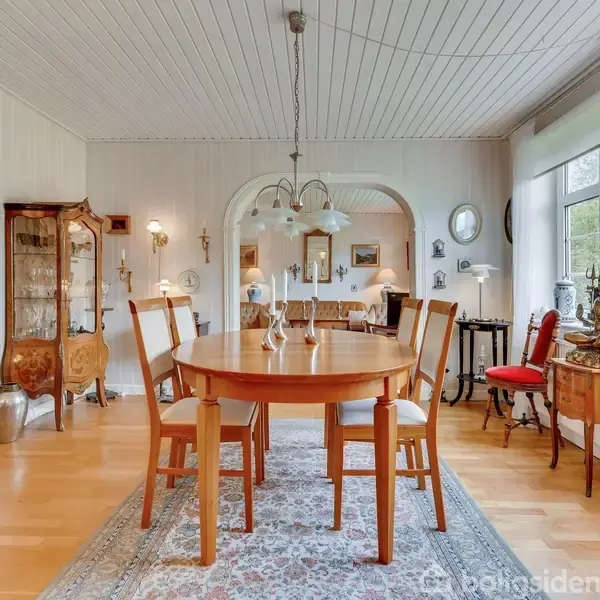 Rundt spisebord med fem stole i en elegant spisestue med lyse trægulve, tæppe, vitrineskab og buet åbning til en stue. Store vinduer lader naturligt lys ind.