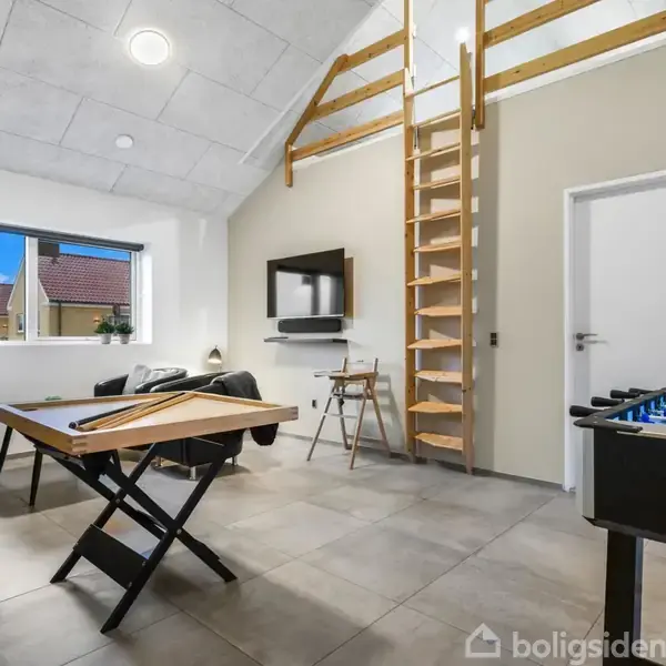 En stue med en loftstige fører op til en hems. Billiardbordet står i midten, omgivet af sofaer. Et bordfodboldspil står til højre. Vinduet giver dagslys.