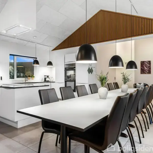 Langt spisebord med sorte stole og hvide pendellamper hængende over. Køkkenø i baggrunden med hvide skabe og indbygget ovn. Moderne og stilrent rum med minimalistisk indretning.