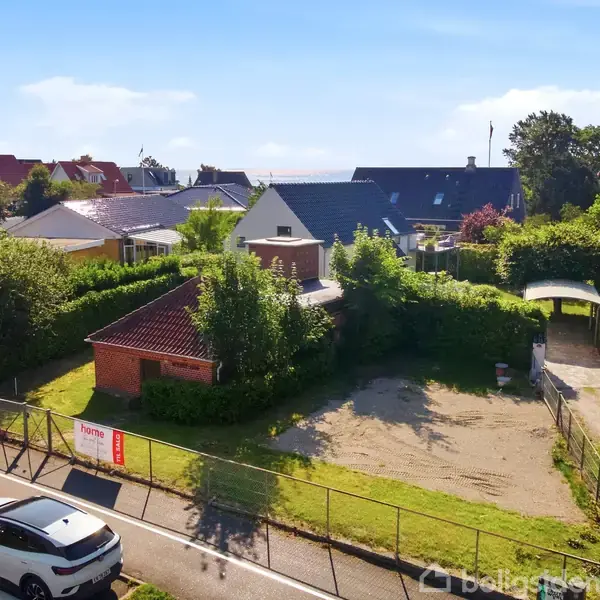 Et fritliggende hus står stille omgivet af grønne hække og naboer. Foran huset ses en parkeringsplads og en til salg-plakat ved vejen. En bil er parkeret udenfor.